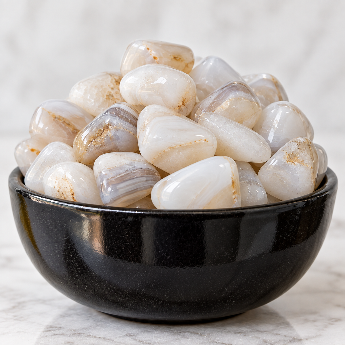 A pile of polished tumbled gemstones in off-white, cream, and light gray with subtle brown veining, displayed in a glossy black ceramic bowl on a light marble surface.