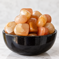 A pile of polished tumbled stones in peach, orange, and translucent cream tones, displayed in a glossy black ceramic bowl on a light marble surface.